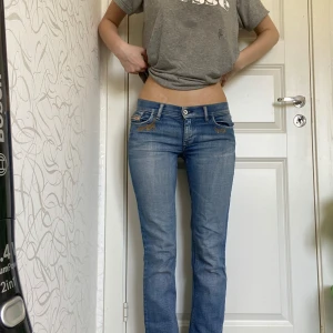 Diesel LIV ljusblå jeans med brodyr med låg midja - Säljer ett par ljusblå Diesel LIV jeans med snyggt broderade detaljer i guld på fickorna. Jeansen har låg midja, raka ben och klassisk femficksdesign. Stretchigt denimtyg för skön passform. Perfekta för dig som gillar en avslappnad men ändå stilren look.