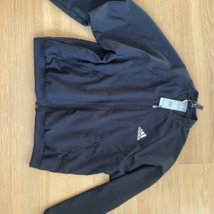 Svart vindjacka från Adidas - Svart vindjacka från Adidas med klassisk logga på bröstet. Jackan har hel dragkedja, två fickor med dragkedja och ribbade muddar. Tillverkad i lätt polyester som andas, perfekt för aktiva dagar. Enkel och stilren design som funkar till det mesta. Priset är ej hugget i sten 