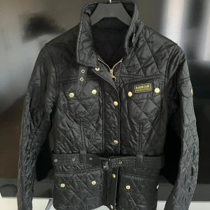Svart quiltad jacka från Barbour Stl 34 - Snygg svart quiltad jacka från Barbour. Stl 34 International med guldiga knappar, bälte i midjan och dragkedja. Jackan har flera fickor och en klassisk Barbour-logga på bröstet. Perfekt för dig som vill ha en stilren och tidlös look.