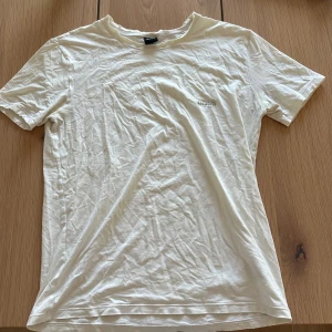 Vit t-shirt från Hugo Boss - Vit t-shirt från Hugo Boss med diskret broderad logga på bröstet. Klassisk rund hals och korta ärmar, tillverkad i mjuk bomull. Enkel och stilren design som funkar till jeans eller shorts. Perfekt för dig som gillar cleana och snygga plagg.
