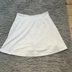 Vit sportig kortkjol med shorts - Säljer en vit sportig kortkjol med inbyggda shorts  med fickor. Kjolen har en enkel och clean design med bred linning och är gjord i ett mjukt, stretchigt syntetmaterial som passar perfekt för aktiva dagar. Superskön och lätt att röra sig i!