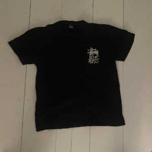 Svart Stüssy t-shirt  - Svart t-shirt från Stüssy i bomull med coolt tryck fram och bak. På bröstet står det 'Stüssy Big and Meaty' i vitt och på ryggen finns en stor dinosaurie i grå toner. Klassisk passform och korta ärmar, perfekt för dig som gillar streetwear.