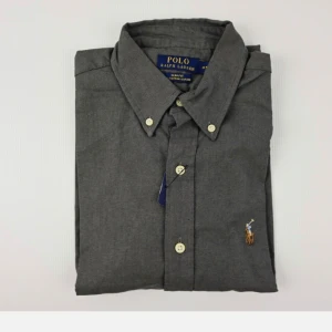 Mörkgrå skjorta från Polo Ralph Lauren - Stilren mörkgrå skjorta från Polo Ralph Lauren med klassisk button-down krage och broderad logga på bröstet. Skjortan har smal passform, vita knappar och är tillverkad i mjuk bomull. Perfekt för dig som gillar en clean och tidlös look.