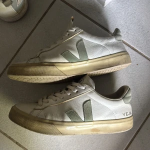 Vita Veja sneakers med gröna detaljer - Snygga vita sneakers från Veja med gröna detaljer och klassisk logga på sidan. Skorna har snörning, platt sula och är tillverkade i skinn med mockadetaljer på hälen. Perfekta för en clean och stilren look.