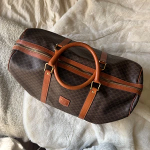 Céline Vintage Macadam Boston Bag - Autentisk Celine Paris väska, tillverkad i Italien. Väskan har det klassiska Celine-mönstret och detaljer i läder. Perfekt för resor eller som en rymlig vardagsväska.   Höjd: 25.5 cm Längd: 49.5 cm Djup: 23 cm