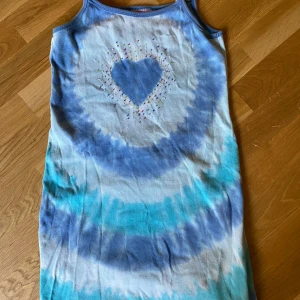 Blå tie-dye klänning med hjärta - Söt kortklänning i blå och vita tie-dye nyanser med broderat hjärta framtill. Klänningen är ärmlös med smala axelband och har ett avslappnat snitt, perfekt för varma dagar. Materialet känns mjukt och skönt mot huden.