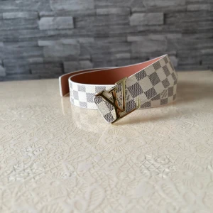 Louis Vuitton rutig skinnbälte - Snyggt Louis Vuitton bälte i skinn med klassiskt rutigt mönster i vitt och grått. Bältet har en ikonisk LV-spänne i guld och detaljerad Louis Vuitton-gravyr på insidan. Perfekt accessoar för att lyfta din outfit med lyxig känsla.