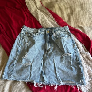 Ljusblå jeanskjol från &Denim - Säljer en ljusblå jeanskjol från &Denim i klassisk femficksmodell. Kjolen har slitna detaljer och fransad nederkant för en cool vibe. Passformen är normal och materialet är robust denim. Perfekt för dig som gillar avslappnad stil.