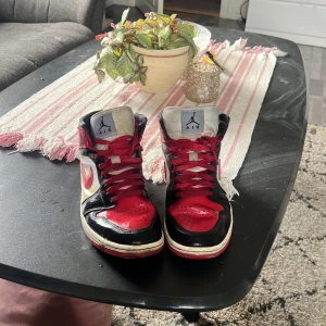 Nike Air Jordan 1 High röd/svart/vit - Säljer ett par ikoniska Nike Air Jordan 1 High med klassisk färgblockning i rött, svart och vitt. Skorna har snörning, rund tå och platt sula. Swoosh-loggan på sidan och Air Jordan Wings vid ankeln. Tillverkade i skinn med röda skosnören för extra streetkänsla.