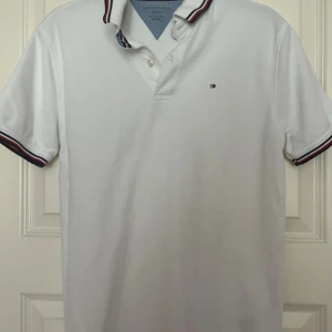Vit pikétröja Tommy Hilfiger XL - Vit pikétröja från Tommy Hilfiger i storlek XL. Klassisk design med korta ärmar och krage med snygga ränder i rött, blått och vitt. Liten Hilfiger-logga på bröstet och knappar vid halsen. Perfekt för en clean och sportig look.