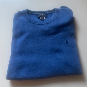 Blå stickad tröja från Polo Ralph Lauren - Snygg blå stickad tröja från Polo Ralph Lauren med klassisk rund halsringning och diskret broderad logga på bröstet. Tröjan har långa ärmar och ribbade muddar vid ärmslut och nederkant. Perfekt för dig som gillar stilren och tidlös design.
