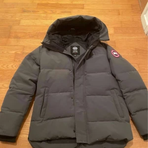 Grå dunjacka från Canada Goose M - Säljer en grå dunjacka från Canada Goose i storlek M. Jackan har en stor huva, quiltad design och klassisk logga på ärmen. 