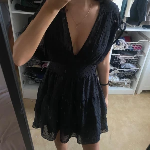 Svart prickig kort klänning från Zara - Med lappen kvar Supersöt svart kort klänning från Zara med djup v-ringning och omlottliv. Klänningen har ett prickigt mönster, volanger i kjolen och är ärmlös. Perfekt för dig som gillar en feminin och trendig look. Materialet är lätt och luftigt, vilket gör den skön att bära.