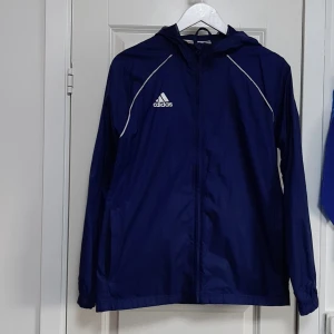 Marinblå vindjacka från Adidas - Snygg marinblå vindjacka från Adidas med vit logga på bröstet och vita detaljer längs axlarna. Jackan har huva, dragkedja framtill och är tillverkad i lätt polyester. Perfekt för dig som vill ha en sportig look och gillar klassisk Adidas-stil.