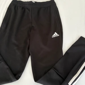 Svarta Adidas mjukisbyxor med vita ränder - Svarta mjukisbyxor från Adidas med klassiska vita ränder längs benen och Adidas-logga på låret. Byxorna har resår i midjan och är gjorda i mjukt material som passar perfekt för chill eller träning.