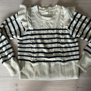 Randig stickad tröja med volang - Superfin stickad tröja från Holly & Whyte by Lindex i storlek S. Den är vit med svarta ränder, har snygga kabelstickade detaljer och volanger vid axlarna. Tröjan har rund hals och långa ärmar med breda muddar. Den har några fläckar på baksidan utav västerarmen.