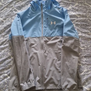 Under Armour blå och grå vindjacka M - Snygg vindjacka från Under Armour i ljusblått och grått. Jackan har huva, dragkedja hela vägen och en bröstficka med dragkedja. Perfekt för dig som gillar sportig stil och vill ha något lätt och funktionellt. Den är slutsåld överallt. Kostar 1200 normalt pris. Skick 9/10. Skulle säga 900kr men kan gå ner i pris vid snabb affär. Den är inte använd så mycket. Passar M och S. Kan tänka mig byta mot nåt också 