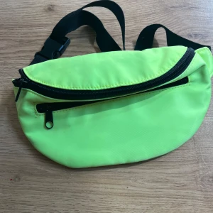 Neongul magväska med svarta detaljer - Säljer en neongul magväska med svarta dragkedjor och justerbart band. Väskan har två fack med dragkedja och är perfekt för att bära det viktigaste. Tillverkad i lätt syntetmaterial och har en clean, sportig look.
