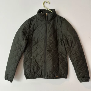 Quiltad grön jacka från Marine - Quiltad mörkgrön jacka från Marine med dragkedja framtill och hög krage. Jackan har lång ärm, elastisk nederkant och diskret logga vid fickan. Perfekt för kyliga dagar och enkel att matcha med olika outfits.