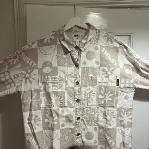 Beige och vit overshirt Smiley x H&M - Overshirt i beige och vitt från Smiley x H&M med coolt rutigt mönster, smileys och blommor. Jackan har två bröstfickor, knappar med smiley-detaljer och är långärmad. Perfekt för dig som gillar statement-plagg och streetstyle.