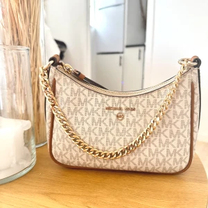 Michael Kors beige axelväska med kedja - Snygg axelväska från Michael Kors i beige och brunt med MK-logga över hela väskan. Väskan har guldfärgad kedja som axelrem och detaljer i brunt skinn. Perfekt storlek för det viktigaste och stängs med dragkedja upptill.