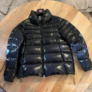 Svart pufferjacka från Colmar - Snygg svart pufferjacka från Colmar med glansig finish och hög krage. Jackan har dragkedja framtill, justerbar nederkant med snörning och Colmar-logga på ärmen. Perfekt för kalla dagar när du vill hålla stilen och värmen.