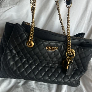 Svart quiltad handväska från Guess - Snygg svart handväska från Guess med quiltat mönster och guldfärgade kedjor. Väskan har flera fack med dragkedja och dekorativa detaljer som hängande berlocker. Tillverkad i skinnimitation och har en lyxig känsla med klassisk design.