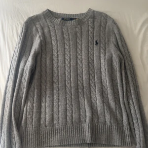 Grå kabelstickad tröja Polo Ralph Lauren - Kabelstickad grå tröja från Polo Ralph Lauren med rund halsringning och broderad marinblå logga på bröstet. Tröjan har långa ärmar och ribbade muddar vid ärmslut och nederkant. Perfekt för lager-på-lager under kyliga dagar. Är även i ett väldigt bra skick och är en stilig övertröja nu till hösten 