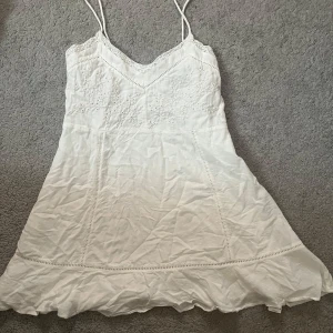 Vit somrig kortklänning med spets - Söt vit kortklänning med tunna axelband och v-ringning. Klänningen har broderade detaljer upptill, spetskant och knappar i ryggen. Volang nertill ger en romantisk känsla. Perfekt för varma dagar och sommarhäng.