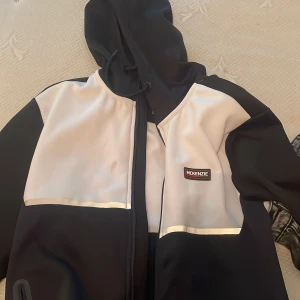 Svart och vit vindjacka från McKenzie - Snygg svart och vit vindjacka från McKenzie med huva och dragkedja framtill. Jackan har två praktiska fickor med dragkedja och coola kontrastpaneler. Perfekt för dig som gillar sportig och stilren look. Märkeslogga på bröstet och justerbar huva med snörning. Fläcken går o ta bort i tväten och tröjan är som m