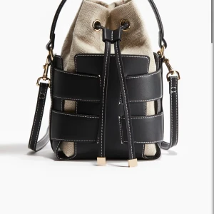 Svart och beige bucket bag med snörning - Cool bucket bag i svart skinn och beige bomull med kontrastsömmar och gulddetaljer. Väskan har snörning upptill och justerbar axelrem. Perfekt för dig som vill ha en unik och trendig accessoar till vardagslooken.