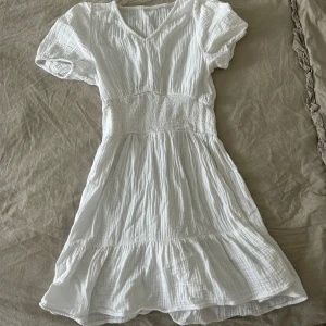 Vit kort sommarklänning med volang - Säljer en vit kort klänning med v-ringning och puffiga korta ärmar. Klänningen har smock i midjan och volang nedtill, vilket ger en flowy och chill vibe. Perfekt för varma dagar och enkel att styla med sneakers eller sandaler.
