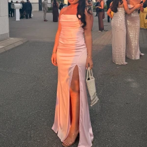 Ljusrosa lång klänning med slits - Säljer en elegant ljusrosa långklänning i satin med axelbandslös design och snygg slits framtill. Klänningen har draperade detaljer över bysten och en figurnära passform som ger en lyxig och modern look. Perfekt för bal eller fest! 💖