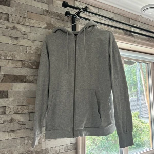 Grå hoodie med dragkedja från 157 - Säljer en enkel grå hoodie från 157 i storlek S. Tröjan har huva med snören, hel dragkedja framtill och två stora fickor. Materialet är mjukt och känns skönt mot huden, perfekt för chill dagar. Långärmad och med ribbade muddar vid ärmslut och nederkant.