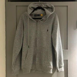Grå hoodie från Polo Ralph Lauren - Snygg grå hoodie från Polo Ralph Lauren med dragkedja och huva. Klassisk logga broderad på bröstet, två fickor fram och ribbade muddar. Perfekt för chill dagar och enkel att matcha med jeans eller joggers. Mjuk bomullsmix som känns skön mot huden.