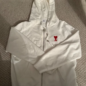 Vit hoodie från Ami med rött hjärta - Säljer en vit hoodie från Ami med dragkedja och snörning vid huvan. På bröstet finns ett broderat rött hjärta med bokstaven A. Tröjan har långa ärmar och är gjord i mjuk bomull, perfekt för chill dagar. !! Har en svart också