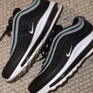 Nike Air Max 97 svart/vit sneakers - Snygga Nike Air Max 97 sneakers i svart med vita detaljer och ikonisk vågig design. Skorna har en synlig Air Max-sula, snörning och reflekterande silvergrå linjer. Perfekta för dig som gillar streetstyle och vill ha en bekväm och trendig sneaker.