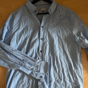 Ljusblå bomullsskjorta  - Snygg ljusblå skjorta från Dressman i mjuk bomull. Klassisk krage, svarta knappar och långa ärmar med dubbla manschettknappar.(kommer strykas innan den skickas)