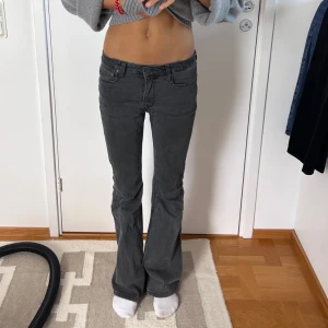 Grå bootcut jeans med låg midja - Säljer ett par grå jeans med snygg bootcut-passform och låg midja. Klassisk femficksmodell med dragkedja och knapp framtill. Jeansen har en mörkgrå ton och är tillverkade i mjukt denimtyg som sitter skönt och ger en cool siluett.
