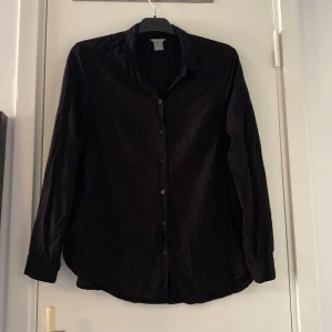 Svart långärmad skjorta - En klassisk svart skjorta med lång ärm och knappar framtill. Skjortan har en enkel krage och rak passform, perfekt att styla till jeans eller kostymbyxor. Materialet känns lätt och smidigt, vilket gör den bekväm att bära hela dagen.