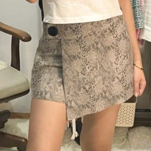Beige omlottkjol med ormmönster - Snygg kort skort i beige med ormmönster och en svart knappdetalj i midjan. Kjolen har en asymmetrisk front och är tillverkad i ett mjukt syntetmaterial.