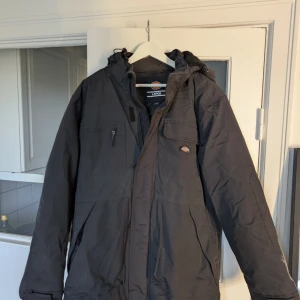 Svart parkajacka från Dickies L - Svart parkajacka från Dickies i storlek L. Jackan har huva, flera praktiska fickor med dragkedja och knapp, samt en klassisk Dickies-logga på bröstet. Perfekt för kalla dagar med sin långa modell och funktionella design.