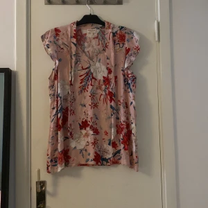 Blommig rosa blus  - Supersöt blommig blus   ljusrosa med röda, blå och vita blommor. Luftig modell med korta ärmar och v-ringad hals. Perfekt för vår och sommar när du vill ha något färgglatt och bekvämt.