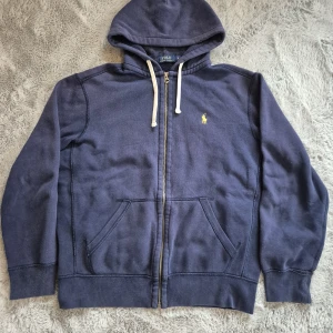 Marinblå hoodie från Polo Ralph Lauren - Snygg och klassisk marinblå huvtröja med dragkedja från Polo Ralph Lauren. Ikoniska broderade loggan i gult på bröstet. Modellen har huva med dragsko och två framfickor. Passar perfekt för både vardagsbruk och en mer avslappnad stil.  Tröjan är i mycket fint skick har alldrig använt de själv. Inga fläckar, hål eller andra defekter.  📦 Kan skickas snabbt och välpackat! ❌ Rök- och djurfritt hem