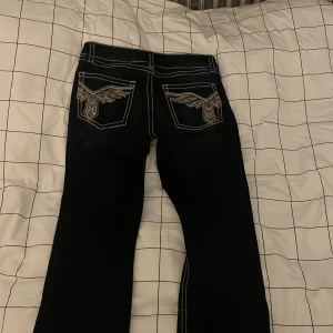 Svarta jeans med broderade fickor Gina Tricot - Svarta jeans från Gina Tricot med kontrasterande vita sömmar och snygga broderade detaljer på bakfickorna. Klassisk femficksmodell i jeansmaterial, perfekt för dig som vill sticka ut lite extra.