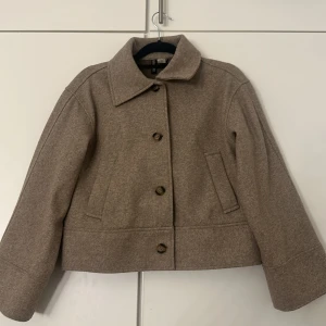 Beige ulljacka med knappar  - Kort beige jacka i ullmix från H&M med klassisk krage och stora knappar framtill. Jackan har raka ärmar, två snedställda fickor och en stilren, minimalistisk look. Perfekt för dig som gillar en clean och tidlös vibe.