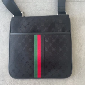 Svart axelväska  - Snygg svart axelväska från ett märke med diskret ett coolt mönster över hela väskan. Framsidan har en ikonisk grön och röd rand, och baksidan har en praktisk dragkedja. Väskan har justerbar axelrem och är tillverkad i slitstark syntet med detaljer i skinn.