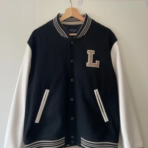 Svart och vit collegejacka Lexington - Collegejacka från Lexington med svart kropp och vita ärmar, broderad L på bröstet och randiga muddar. Jackan har knappar framtill, två fickor och klassisk varsity-stil. Insidan har snyggt Lexington Company-tryck. Perfekt för en sportig och trendig look.