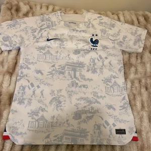 Frankrike Nike fotbollströja mönstrad - Snygg vit Frankrike fotbollströja från Nike med blått tryck och broderat FFF-emblem med tupp och två stjärnor. Tröjan har korta ärmar och ett unikt mönster med franska motiv och flaggor. Materialet är lätt och andas, perfekt för match eller häng.
