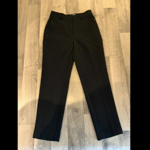 Kostymbyxor  - Klassiska svarta kostymbyxor från NA-KD med raka ben och pressveck. Byxorna har hällor för bälte, sidfickor fram och fuskfickor bak. Perfekta för en clean och stilren look. Materialet känns mjukt och följsamt.